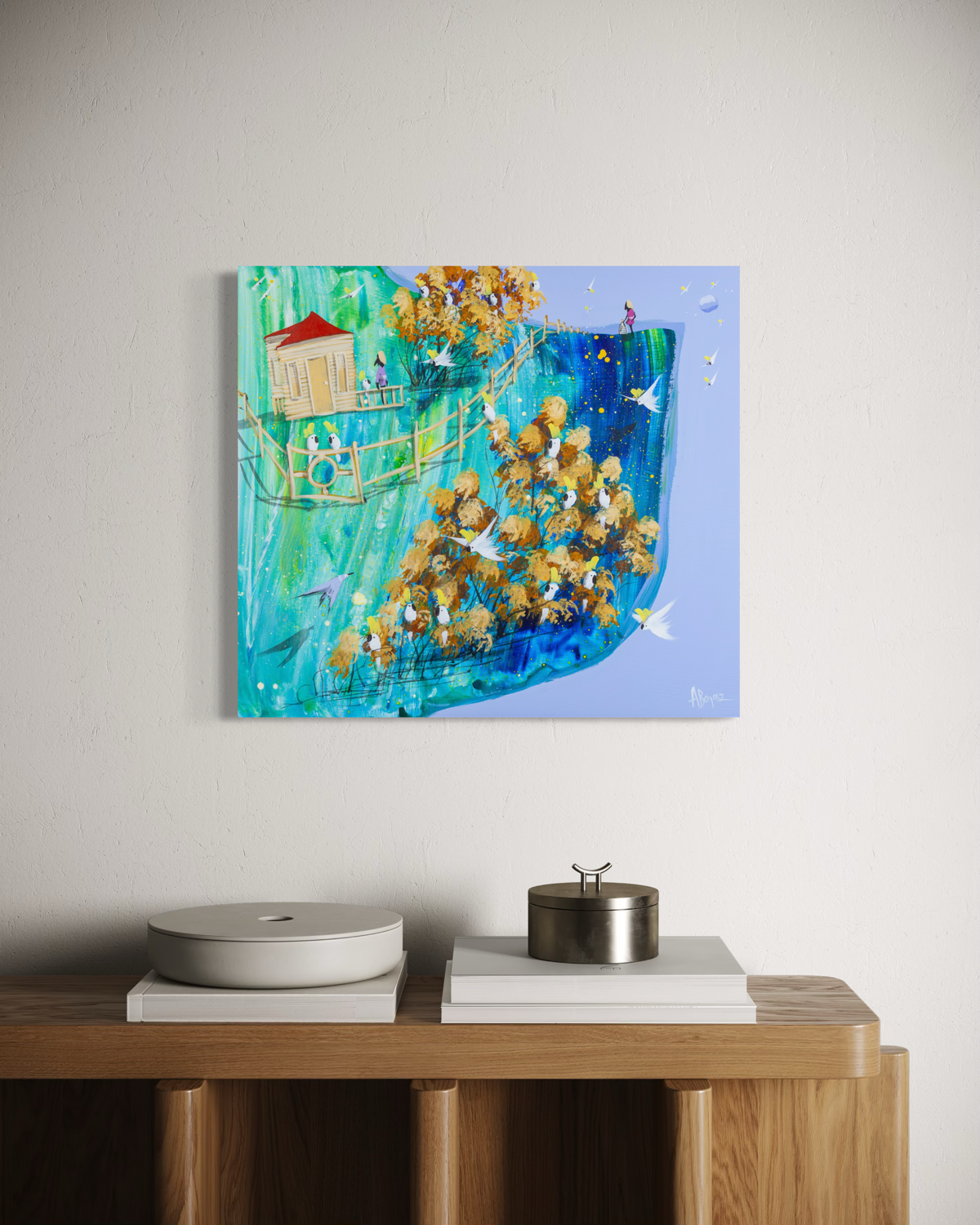 The Open Cage by Adam Bogusz, 50 x 45 cm painting on MDF board hanging above an oak sideboard with white books, a pale ceramic dish and a metal canister, showing a cottage, a long fence and golden gums full of cockatoos.