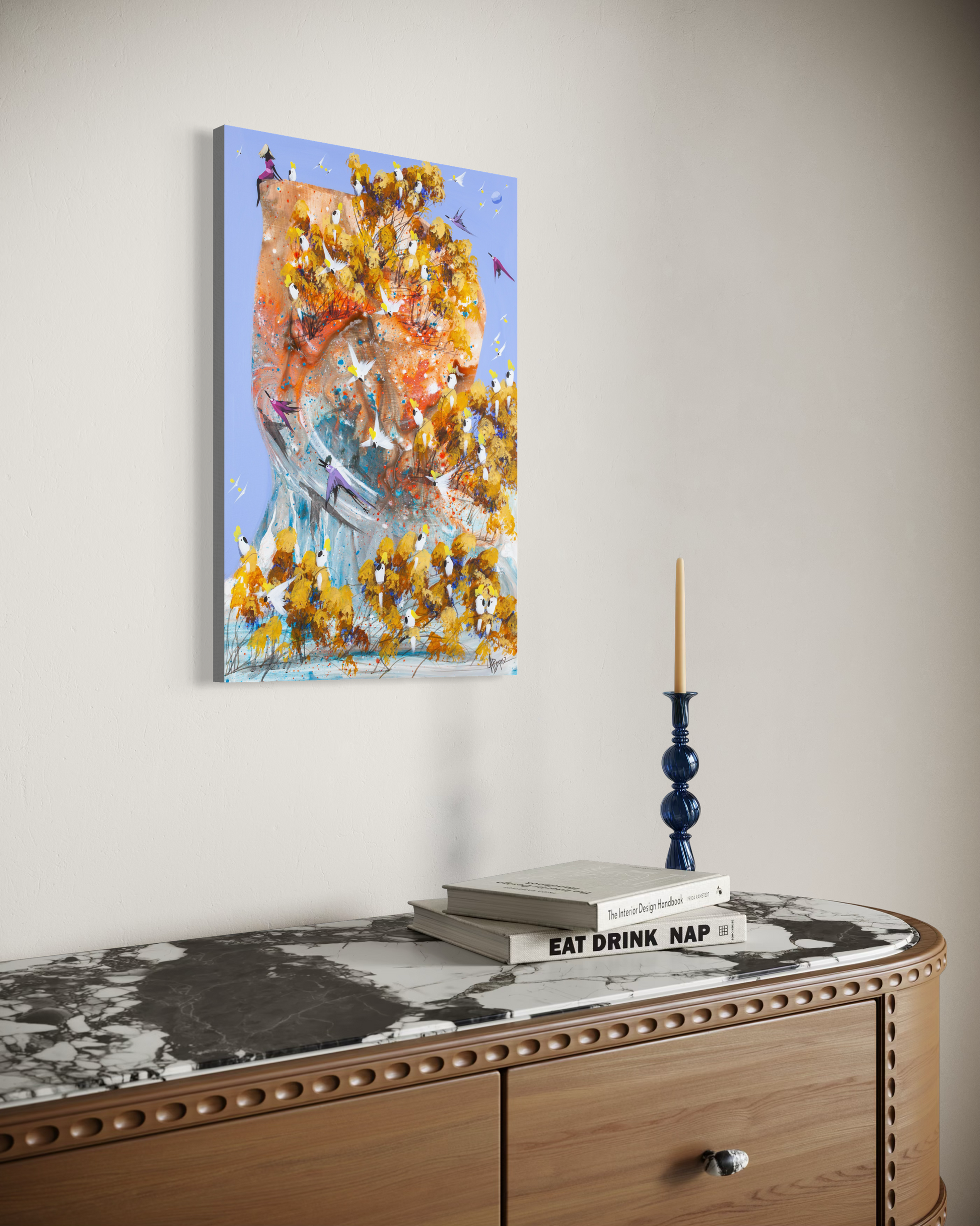 A Good Place to Land by Adam Bogusz displayed on a pale wall above a fluted timber sideboard with a marble top, styled with books and a blue glass candlestick, shown to indicate scale.