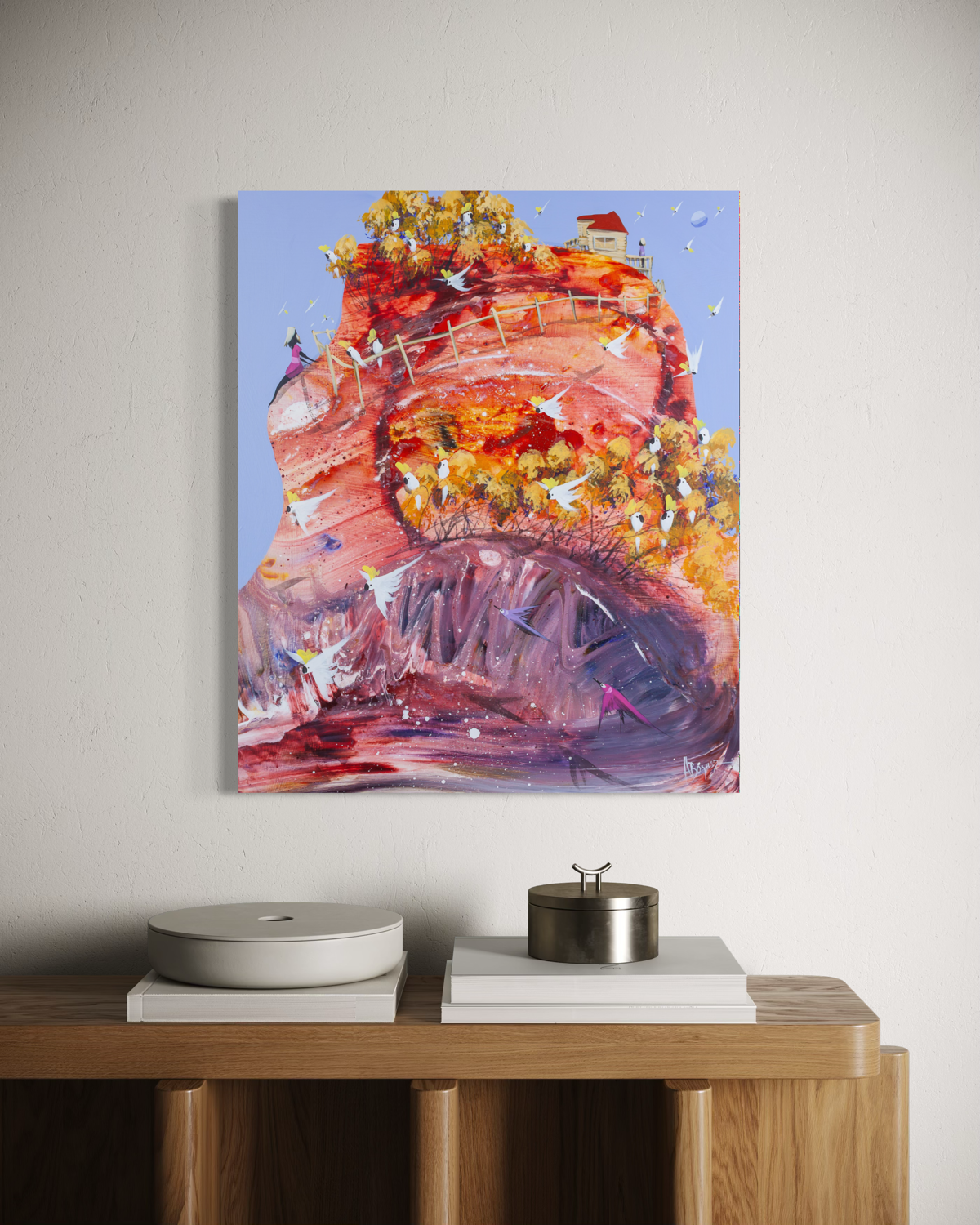 The Wiggly World by Adam Bogusz, 60 x 50 cm painting on MDF board hanging above an oak sideboard with white books, a pale ceramic dish and a metal canister, showing a swirling red and purple hill crowned with golden trees full of cockatoos.