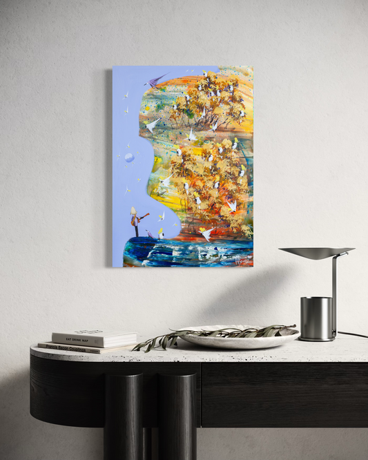 His Own Song by Adam Bogusz, 50 x 35 cm painting on MDF board hanging above a black console with a travertine top, a stone bowl holding an olive branch, books and a metal lamp, showing golden gums full of cockatoos and a small figure playing guitar by the water.