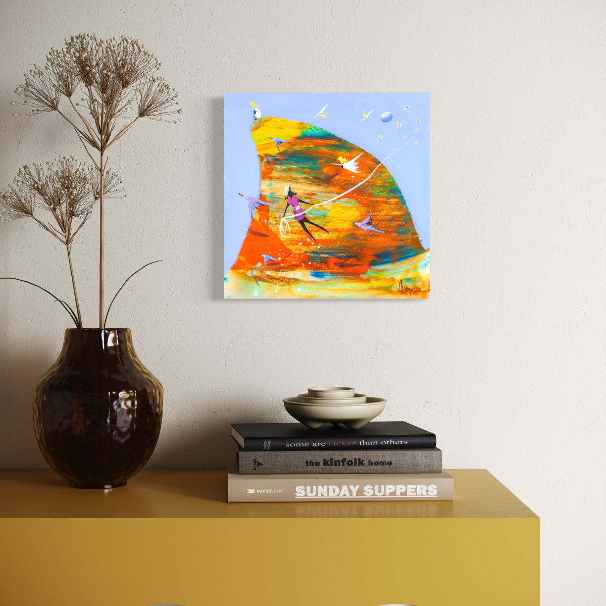 Carried Away by Adam Bogusz hanging above a mustard yellow sideboard with a dark glass vase of dried flowers, stacked books and ceramic bowls, shown to scale in a living space.